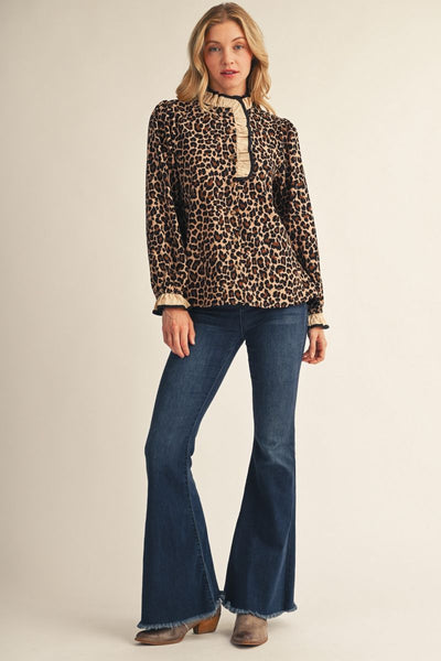 A woman modeling a leopard-print long-sleeve blouse with a high mock neck and beige trim, paired with dark blue flare jeans and brown boots, against a solid light beige studio background.