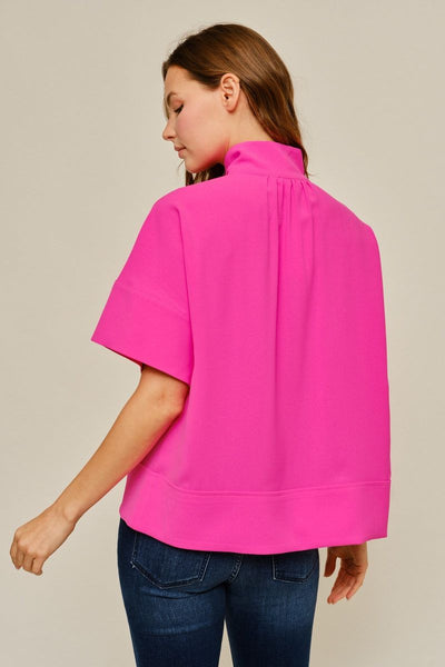 A woman shown from the back wearing a bright hot pink short-sleeve blouse with a gathered high neck, paired with dark blue jeans, against a solid light beige background.