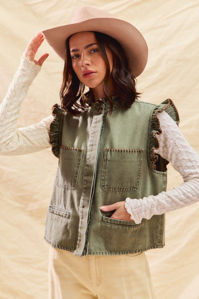 Woman wearing a green vest with ruffled details and a beige hat against a beige background