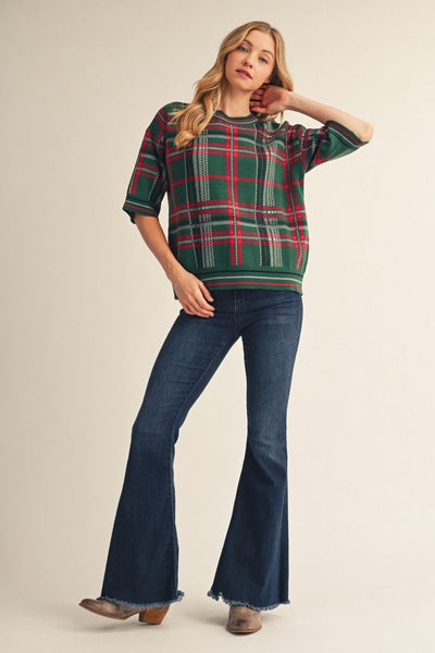 A woman modeling a green, red, and white plaid short-sleeve knit sweater paired with dark blue flare jeans and brown boots, set against a solid light beige studio background.