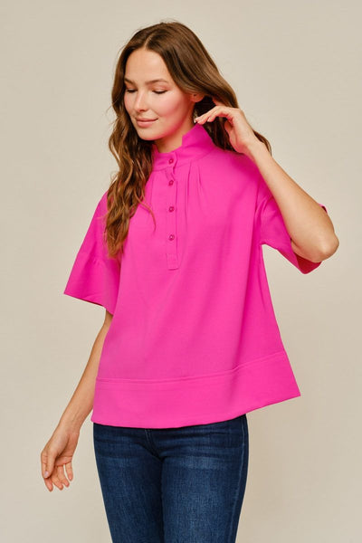 A woman wearing a bright pink short-sleeve mock-neck top with a button placket and blue jeans, standing against a plain beige studio background.