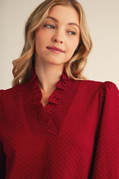 A close-up of a woman wearing a textured deep red blouse with a ruffled smocked neckline and puffed sleeves, set against a plain light beige background.