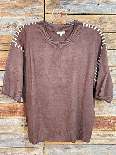 A brown knit sweater with white contrast stitching along the shoulders and sleeves, hanging on a clear plastic hanger against a rustic wooden plank background.