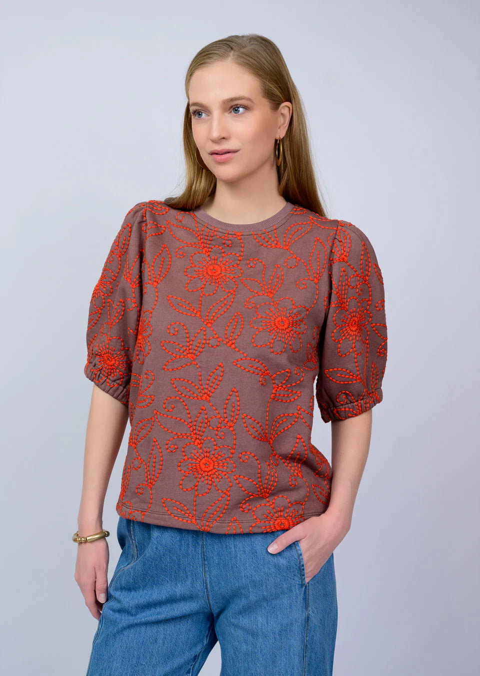 Woman wearing a red and brown patterned blouse with blue jeans on a gray background