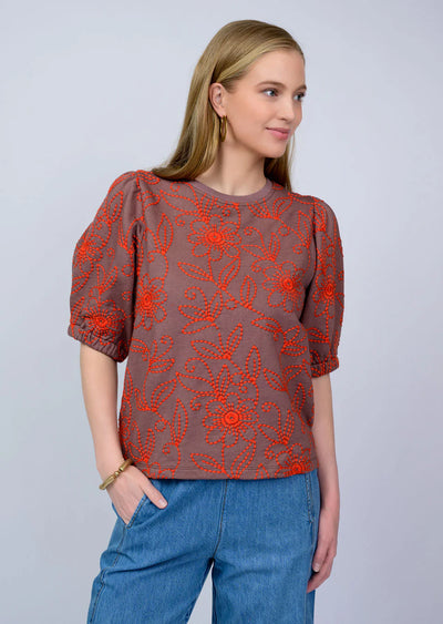 Woman wearing a brown blouse with red floral patterns and blue pants on a light gray background