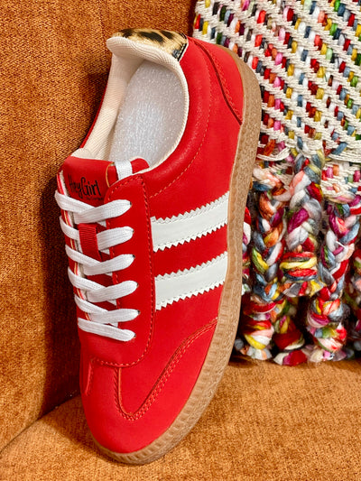 A red leather sneaker with white side stripes, white laces, and a tan rubber sole, photographed leaning against a brown textured surface next to a multicolored woven textile.