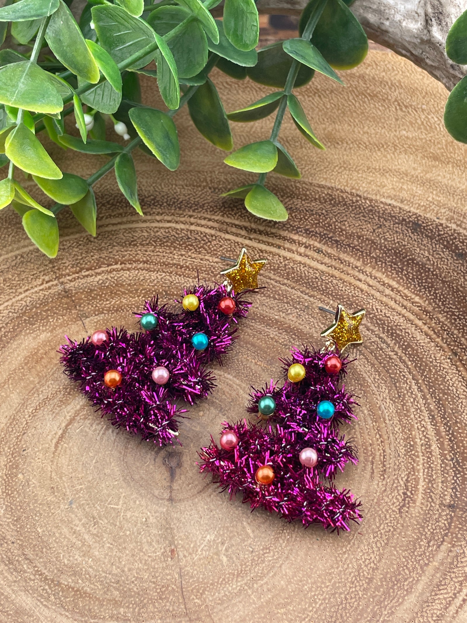 pink tinsel christmas tree earrings with colorful beads on a wooden surface with green leaves.