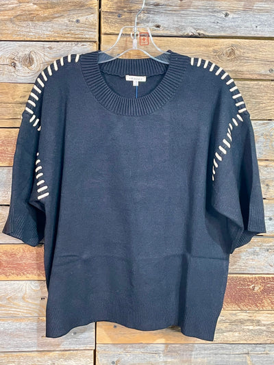 A black knit sweater with white striped detailing on the shoulders and sleeves, hanging on a clear plastic hanger against a rustic wooden plank background.