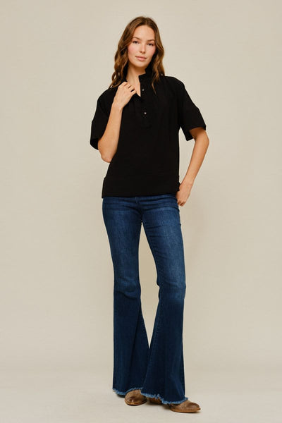 A woman modeling a black short-sleeve polo shirt and dark blue flare jeans with brown shoes, standing against a plain beige studio background.
