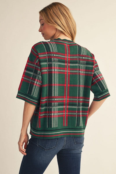 A woman shown from the back wearing a green short-sleeve knit top with a red and white plaid pattern, paired with dark blue jeans, against a solid beige background.