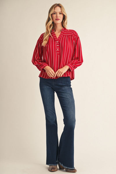 A woman modeling a red and white vertically striped long-sleeve blouse with dark blue flared jeans, standing against a plain beige studio background.
