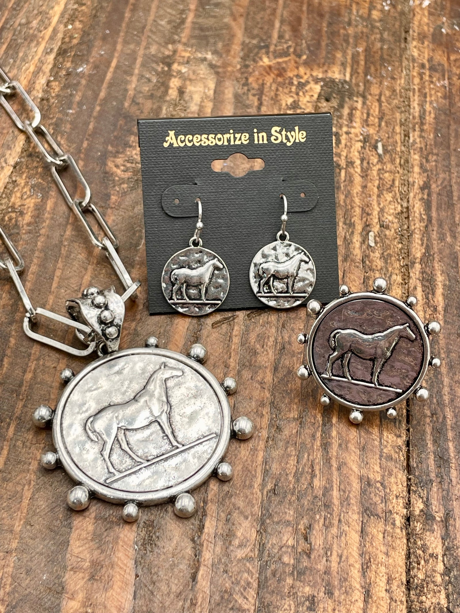 A set of silver-colored jewelry including a paperclip chain with a coin pendant, a ring, and earrings, displayed on a wooden surface with a black background carrying product information.