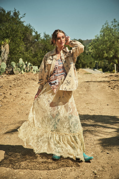 Woman in a long, light-colored dress standing on a dirt road with trees in the background