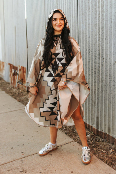 Woman wearing a patterned dress and beige coat standing against a corrugated metal wall.