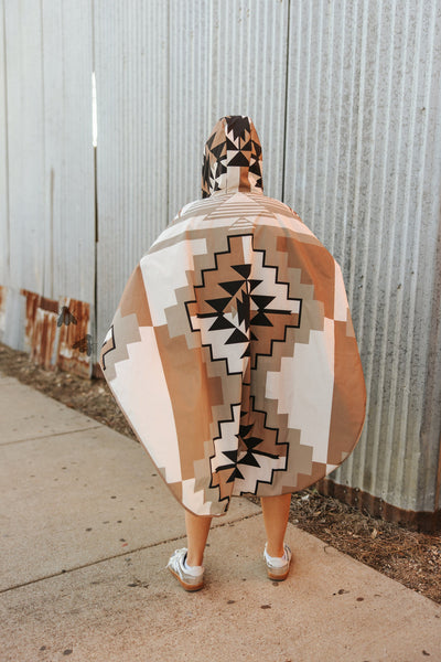 Person wearing a patterned poncho standing on a sidewalk with a corrugated metal wall in the background.