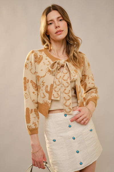 Woman wearing a beige and brown patterned cardigan with a white skirt against a neutral background