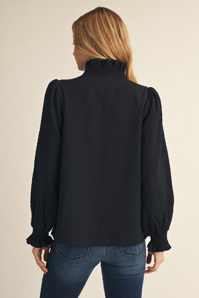 A woman viewed from the back wearing a black textured long-sleeve blouse with a ruffled mock neck and puffed shoulders, paired with blue jeans, against a plain beige background.