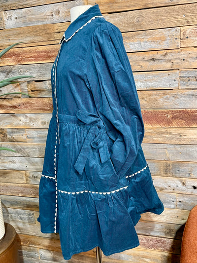 A teal blue velvet long-sleeve blouse with white contrast stitching, displayed on a mannequin torso against a rustic wooden plank wall.