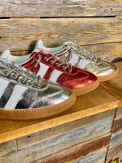 Three metallic low-top sneakers arranged on a wooden surface against a rustic wood plank background. The foreground shoe is metallic silver and white, the middle shoe is metallic red and white, and the rear shoe is metallic gold and white.
