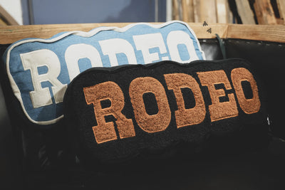 Two doormats with 'RODEO' text, one blue and white, one brown and black, on a wooden surface.