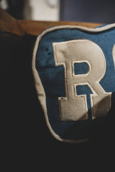 Blue pillow with a large beige letter 'R' on a dark background