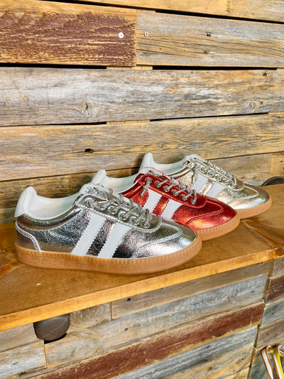 Three metallic low-top sneakers displayed on a wooden shelf against a rustic wood plank wall. The shoes have a silver and red metallic finish with white side stripes and gum soles.