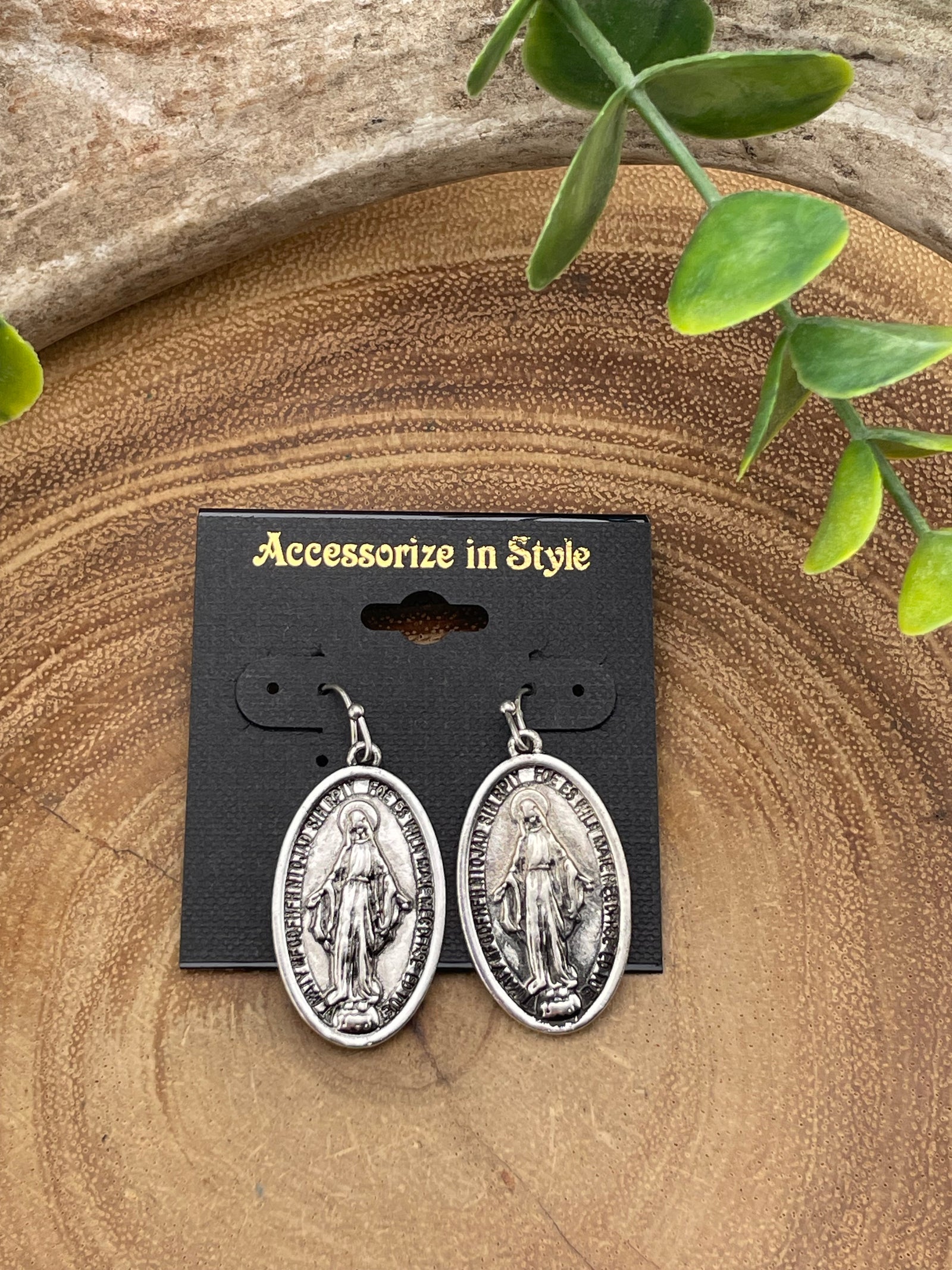 Silver earrings on a black card with 'Accessories in Style' text, placed on a wooden surface with green leaves.