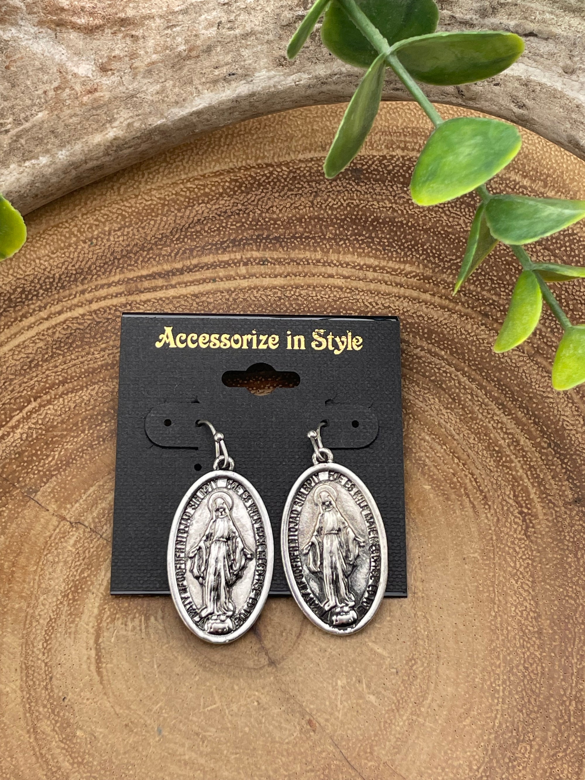 Silver earrings on a black card with 'Accessories in Style' text, placed on a wooden surface with green leaves.