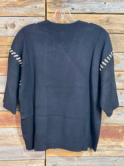 A black knit sweater with white dashed stripe details on the sleeves, hanging on a clear plastic hanger against a rustic wooden plank background.