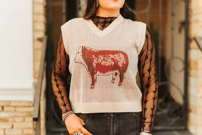 Person wearing a sleeveless sweater with a cow design in front of a brick wall.