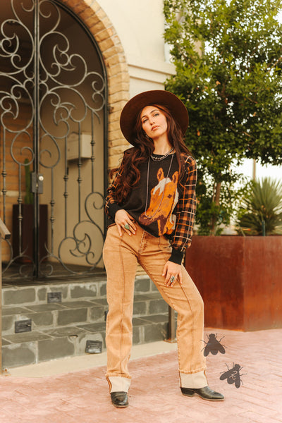 Woman wearing a plaid shirt with a fox design, tan pants, and a hat standing in front of a decorative archway.
