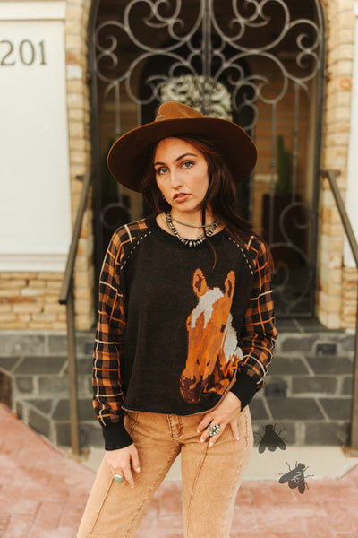 Woman wearing a sweater with a horse design and plaid shirt in front of a building.
