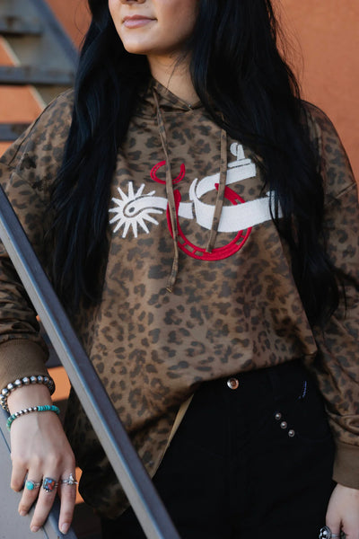 Person wearing a leopard print hoodie with a graphic design, standing against a brown background.