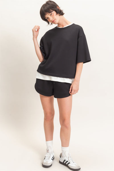model wearing black short sleeve top with white tshirt underneath, black shorts and tennis shoes in front of a light background
