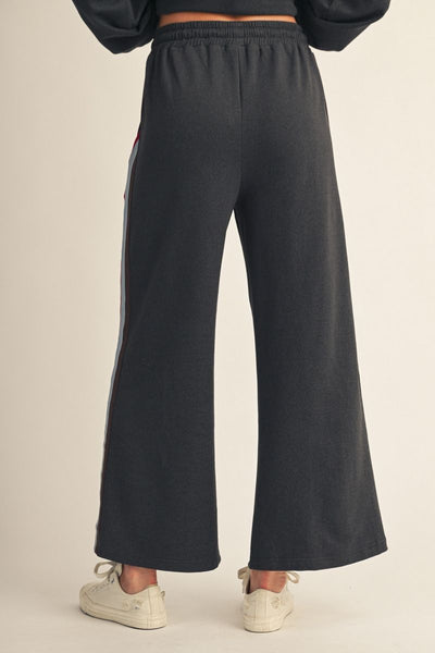 A person shown from the back wearing dark charcoal wide-leg sweatpants with a side stripe detail, paired with white sneakers, against a plain beige studio background.