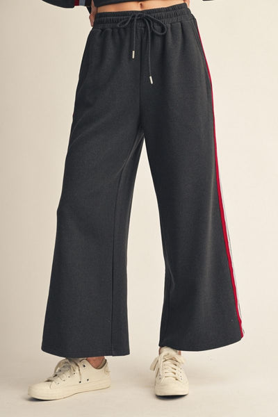 Black wide-leg sweatpants with a red side stripe and white drawstrings, shown on a model wearing white sneakers against a light beige background.