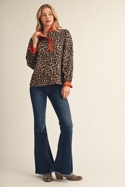 A woman modeling a long-sleeve leopard-print blouse with bright red ruffled trim at the collar and cuffs, paired with dark blue flare jeans and brown boots, against a plain beige studio background.