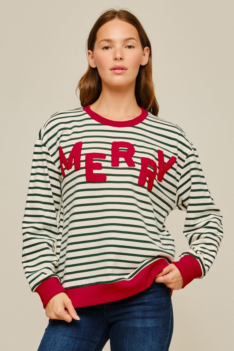 A woman wearing a festive long-sleeve crewneck sweatshirt with green and white horizontal stripes, red ribbed collar, cuffs, and hem, featuring the word 'MERRY' in red across the chest. She is wearing blue jeans against a solid light beige background.