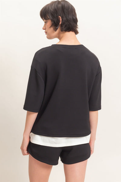 back of model wearing black short sleeve top with white tshirt underneath and black shorts in front of a light background