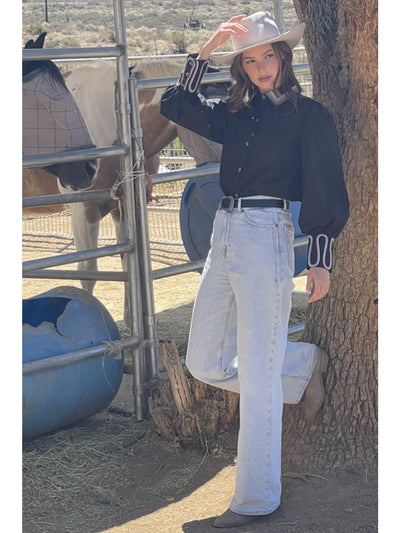 Person wearing a black shirt and white jeans leaning against a tree in a rural setting with a horse in the background.