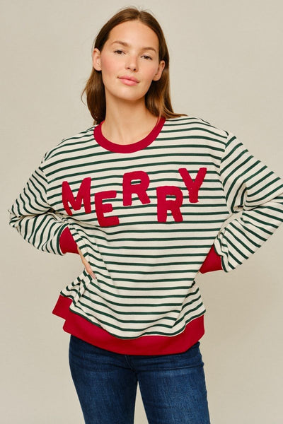 A woman wearing a festive long-sleeve crewneck sweater with green and white horizontal stripes, red ribbed trim, and the word 'MERRY' in large red letters across the chest, paired with blue jeans against a neutral beige background.