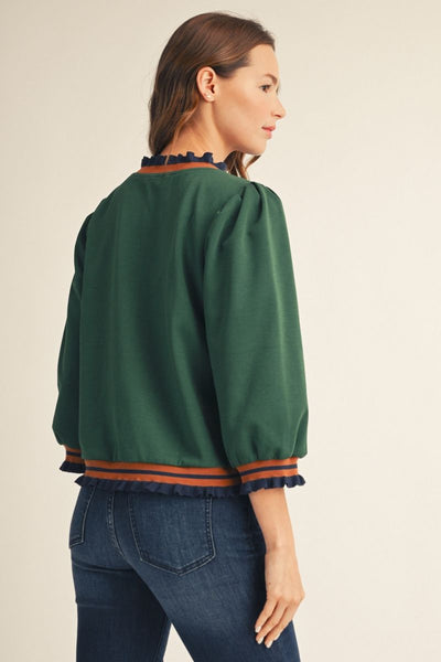 A woman shown from the back wearing a dark green cropped blouse with orange and navy ruffled trim, paired with blue jeans, against a plain beige studio background.