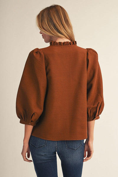 A woman shown from the back wearing a rust-brown long-sleeve blouse with a ruffled high neckline and puffed sleeves, paired with blue jeans, against a plain beige studio background.