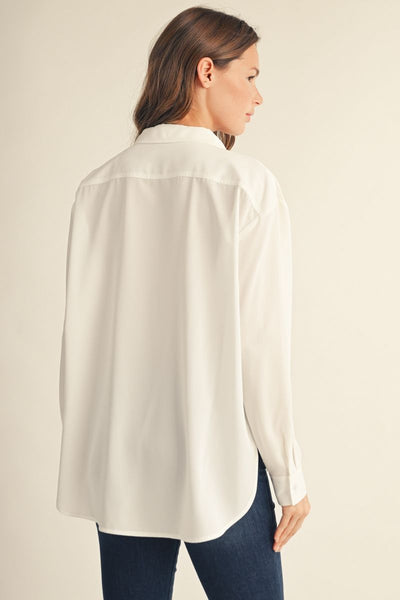 A woman shown from the back wearing an oversized white button-up shirt and dark blue jeans, photographed against a plain light beige studio background.