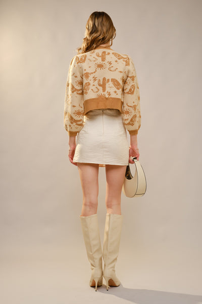Woman wearing a patterned jacket, white skirt, and beige knee-high boots against a plain background