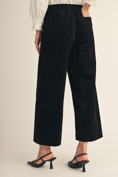 back of model wearing a white blouse, black heels and black corduroy pants on a beige background