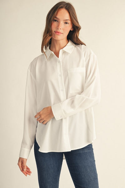 A woman modeling a white long-sleeve button-up shirt with a chest pocket, paired with dark blue jeans, against a plain light beige studio background.