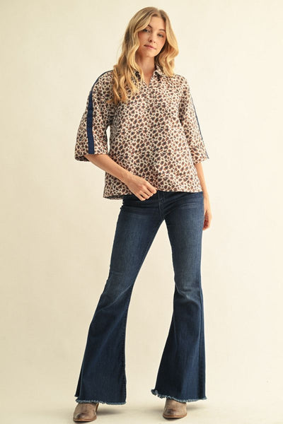 A woman modeling a three-quarter sleeve button-up shirt with a beige and brown leopard print and navy blue side stripes, paired with dark blue flare jeans and brown boots, against a solid light beige background.