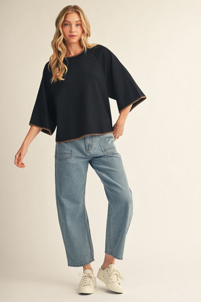 A woman modeling a black short-sleeve top with wide, draped sleeves and light-wash blue cropped jeans, paired with white sneakers, against a plain beige studio background.