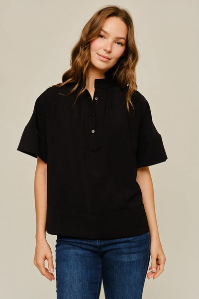 A woman wearing a black short-sleeve polo shirt with a buttoned collar and blue jeans, standing against a plain beige studio background.